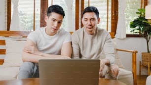 Feliz pareja gay asiática joven se sienta en el sofá usar una videollamada de facetime de computadora portátil con amigos y familiares en la sala de estar de casa.