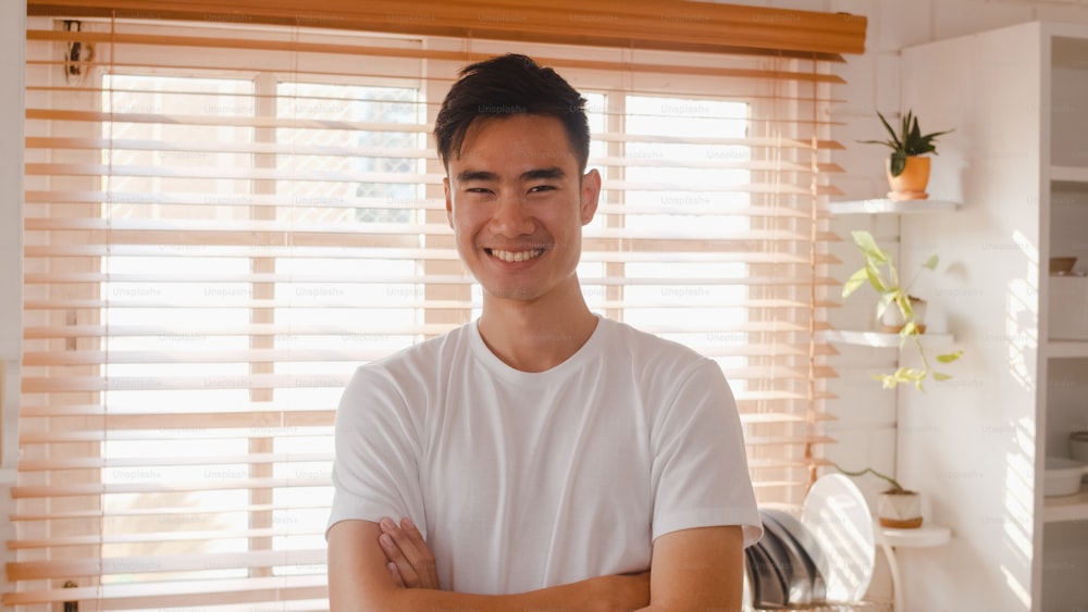 Happy young Asian male feeling happy smiling, arms crossed and looking to camera while relax in kitchen at home.