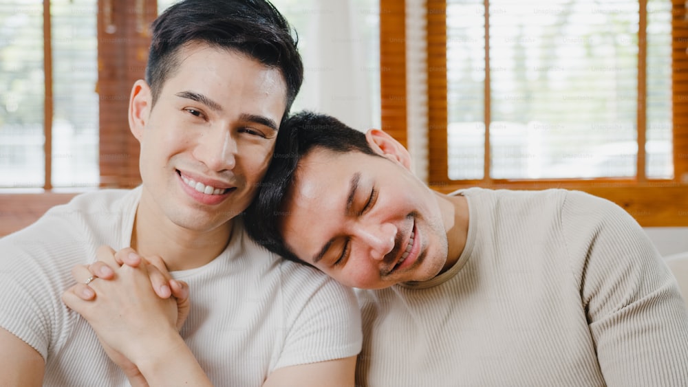 Ritratto Giovane Asia Coppia gay che si sente felice mostrando l'anello a casa. Gli uomini LGBTQ+ dell'Asia si rilassano con un sorriso a denti stretti guardando la telecamera mentre si abbracciano nel moderno soggiorno di casa nel concetto mattutino.