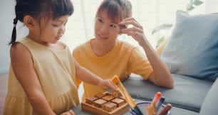 행복한 아시아 가족 엄마는 집 거실의 소파에서 휴식을 취하는 나무 상자로 소녀에게 보드 게임 취미를 가르칩니다. 함께 시간 보내기, 사회적 거리두기, 코로나바이러스 격리.