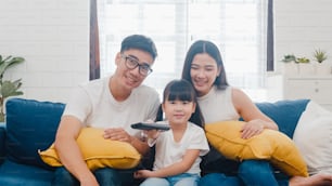 Stile di vita Papà, mamma e figlia coreani che guardano la TV insieme e si divertono sdraiati sul divano nel soggiorno in una casa moderna.