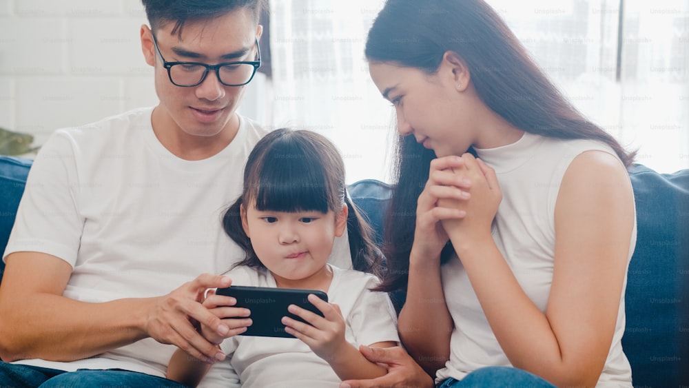 Happy Asian family dad, mom and daughter playing funny game online on smartphone sitting sofa in room at house.