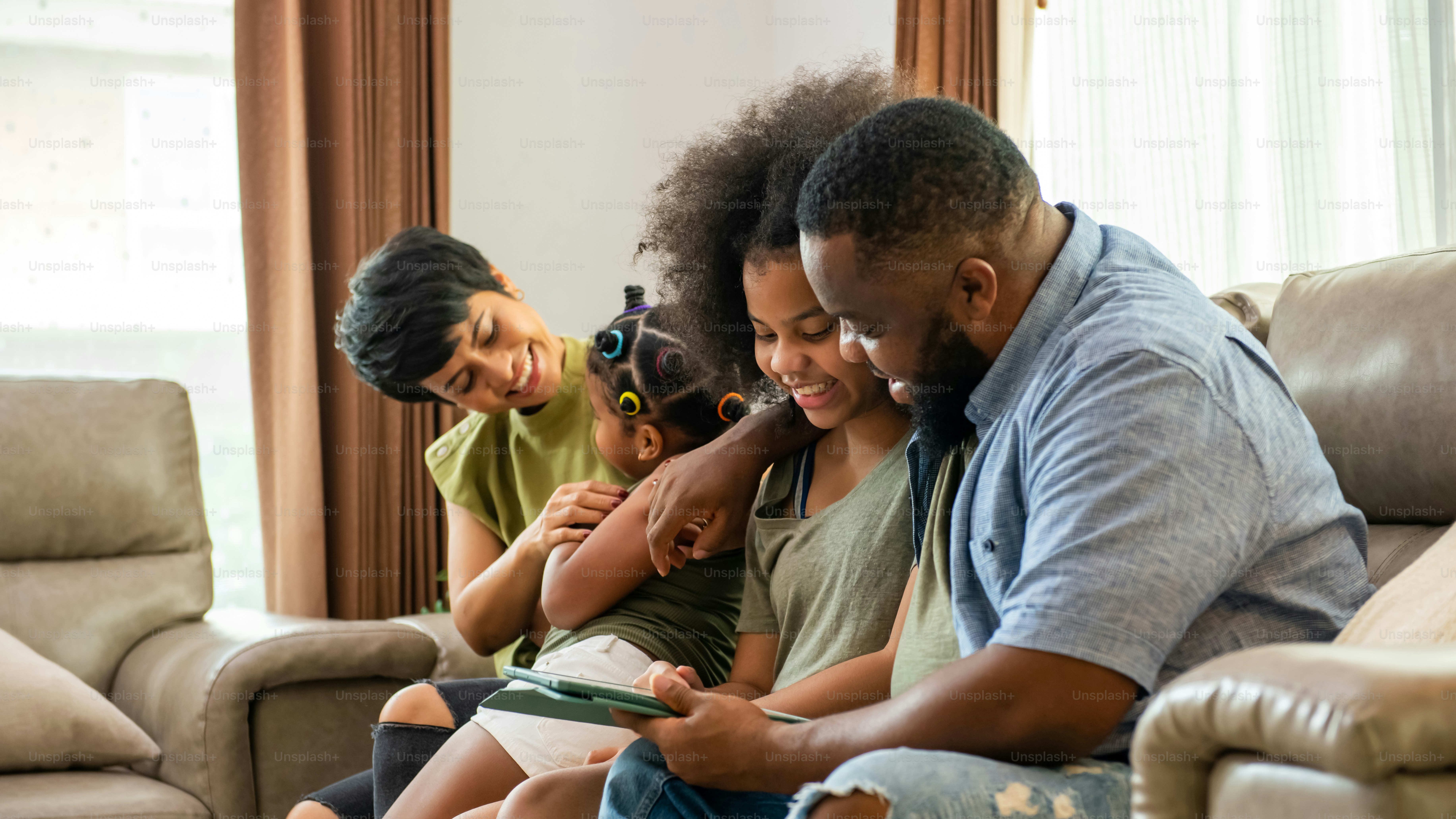 African mixed race parents and two little daughter sitting on sofa in living room using digital tablet play games or watch movie together. Happy family enjoy weekend activity with technology together at home