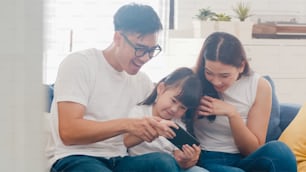 Happy Asian family dad, mom and daughter playing funny game online on smartphone sitting sofa in room at house.