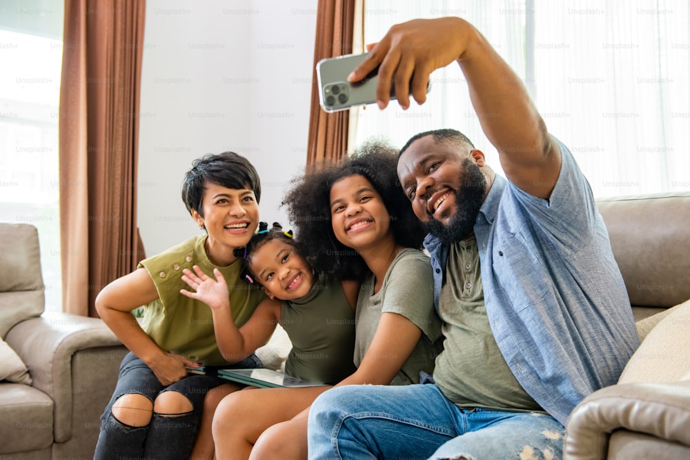 행복한 아프리카 부모와 두 어린 딸이 거실 소파에 앉아 휴대폰을 사용하여 함께 셀카를 찍습니다. 행복한 가족은 집에서 기술로 즐거운 여가 활동을 즐기고 있습니다.