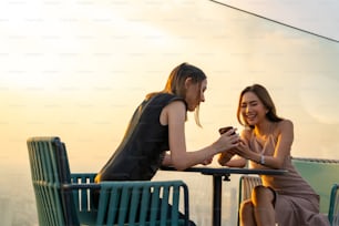 Confiance Amis asiatiques utilisant un smartphone pour les médias sociaux ou les achats en ligne ensemble tout en se rencontrant et en dînant dans un restaurant sur le toit en plein air d’un gratte-ciel dans la ville au coucher du soleil d’été.