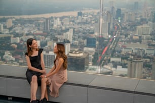 Beautiful Asian woman friends meeting and talking together at skyscraper rooftop restaurant in metropolis at summer sunset. Smiling female friend enjoy outdoor lifestyle activity in the city at night
