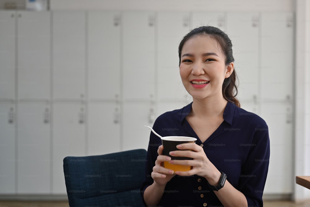 Cheerful asian woman holding cup of instant noodle and smiling to camera.