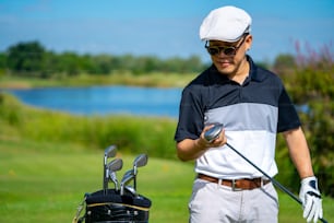 Portrait d’un homme asiatique souriant tenant un club de golf debout sur le fairway du terrain de golf par une journée ensoleillée. Un golfeur masculin en bonne santé profite d’une activité de plein air en jouant au golf au country club pendant les vacances d’été
