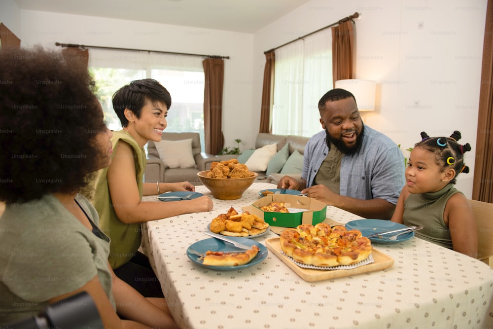 Heureux parents de famille africains et deux petites filles mangeant du poulet frit et de la pizza pour le dîner ensemble. Père et mère et jolie petite fille s'amusent et s'amusent à manger et à partager un repas ensemble à la maison