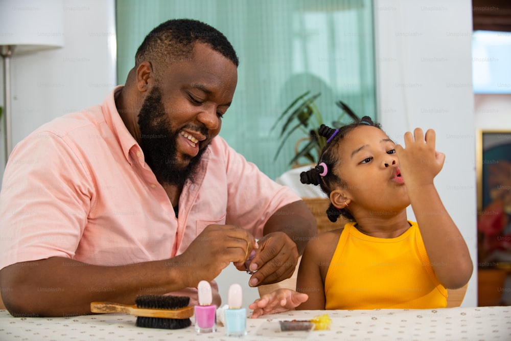 Happy family African father applying nail polish to little daughter in living room. Dad and child girl kid enjoy and having fun leisure activity spending time together with beauty treatment at home
