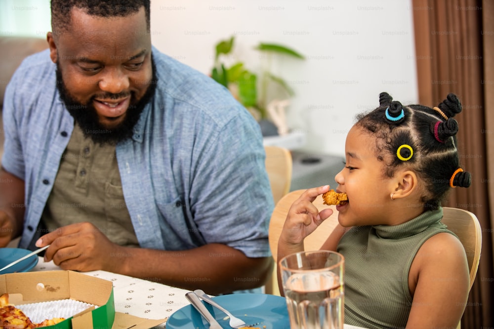 Happy African family parents and two little daughter eating fried chicken and pizza for dinner together. Father and mother and cute child girl kid enjoy and fun eating and sharing a meal together at home