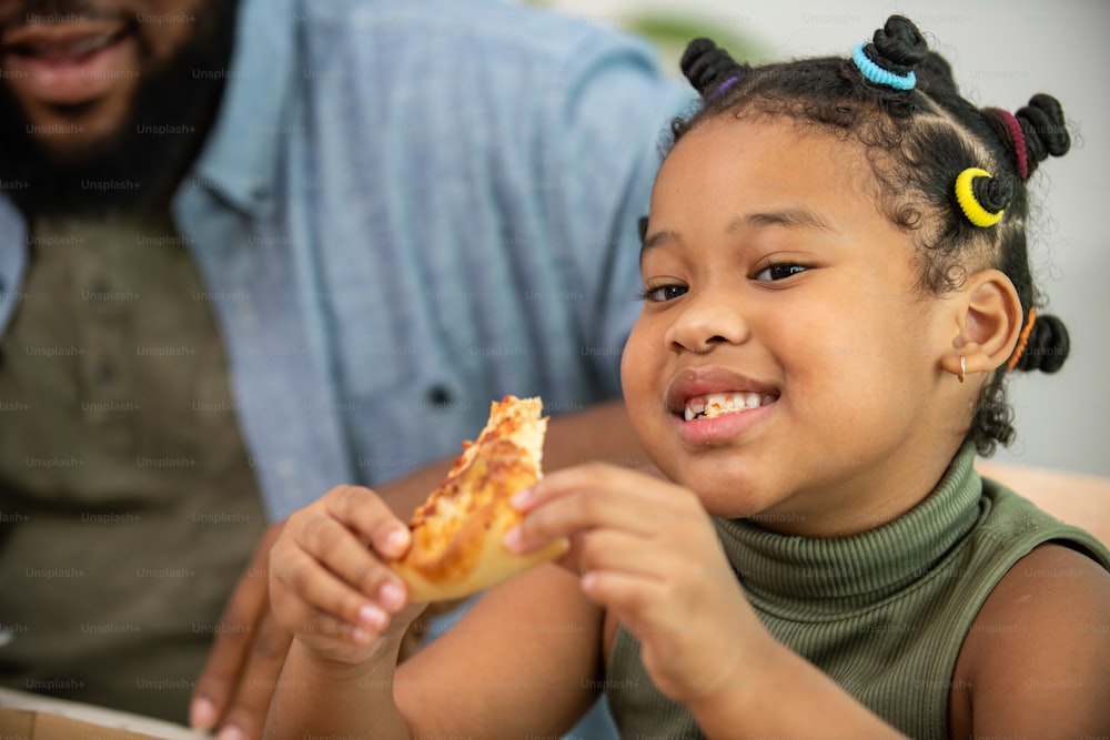 Des parents heureux d’une famille africaine et deux petites filles mangent du poulet frit et de la pizza pour le dîner ensemble. Le père et la mère et l’enfant mignon fille l’enfant apprécient et s’amusent à manger et à partager un repas ensemble à la maison