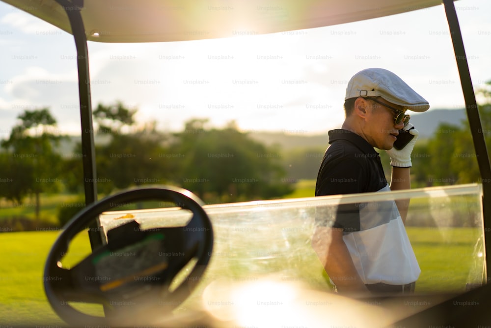 자신감 아시아 사업가는 컨트리 클럽에서 골프를 치는 동안 휴대 전화로 일하는 기업 비즈니스로 이야기하면서 골프 카트에 앉아 있습니다. 건강한 남성 골퍼는 여름 방학에 야외 스포츠를 즐깁니다.
