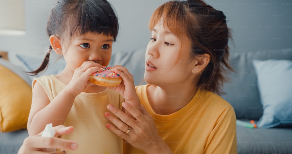 Happy Asia Familie Mutter und Kleinkind Mädchen Essen Donuts und Spaß entspannen genießen auf der Couch im Wohnzimmer im Haus. Zeit miteinander verbringen, soziale Distanz, Quarantäne für Coronavirus.