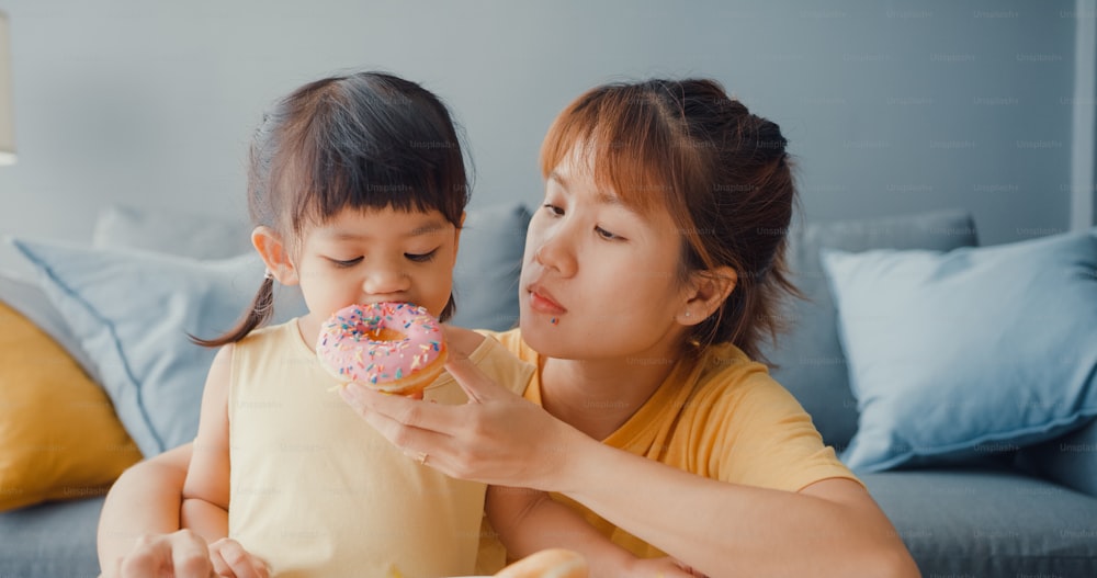 Happy Asia family mom and toddler girl eating donuts and having fun relax enjoy on couch in living room at house. Spending time together, Social distance, Quarantine for coronavirus.