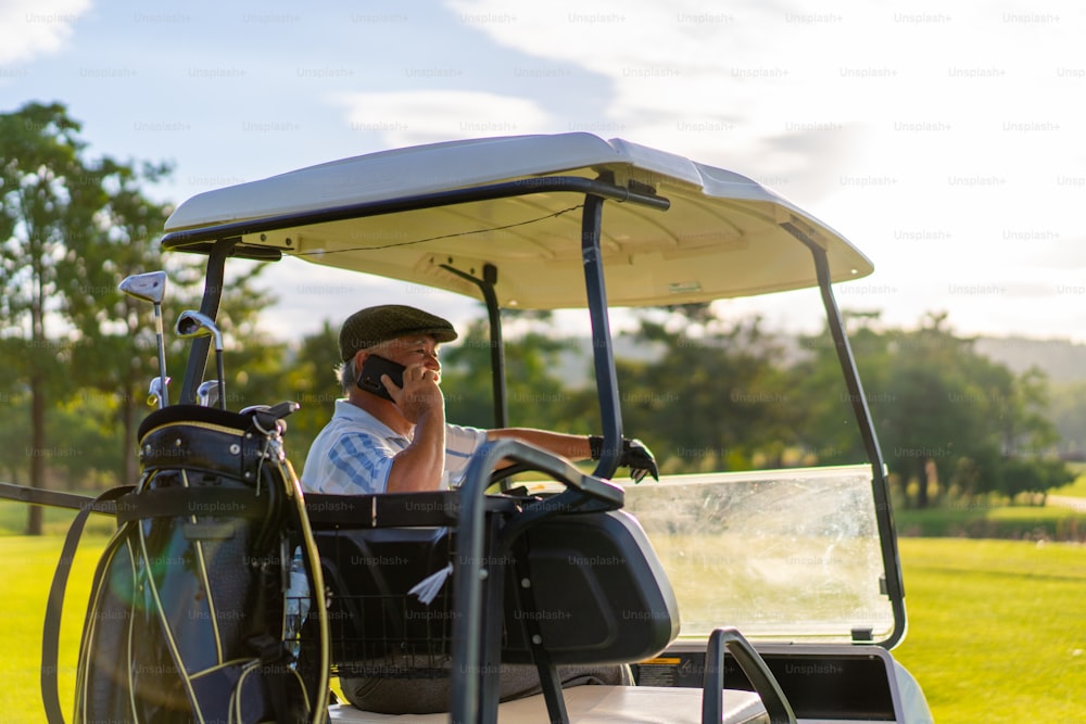 Uomo d'affari anziano asiatico seduto sul carrello da golf con parlare al telefono cellulare che lavora affari aziendali mentre gioca a golf con al country club. L'anziano golfista maschio si diverte con lo sport all'aria aperta durante le vacanze estive.