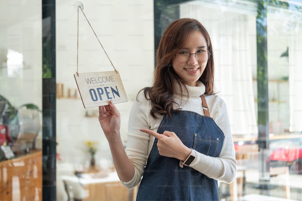 笑顔でコーヒーショップでカメラを見ているポートレートアジアの女性。開いた看板を手にしたバリスタの女性。働く女性の中小企業経営者または中小企業のコンセプト。