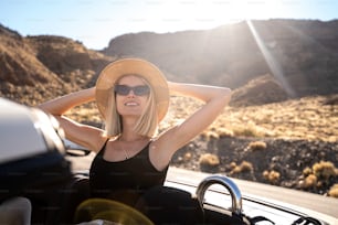 Smiling blonde caucasian woman wearing hat and sunglasses enjoying her summer road trip in cabrio . Freedom. Travel. Tourism. Vacation.