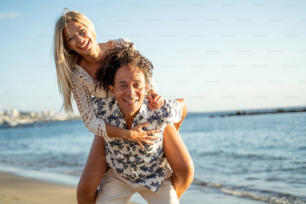 Smiling happy multiethnic couple having fun together on the beach, during sunset, looking to the camera. A lot of copy space. Real people emotions. Summer time.