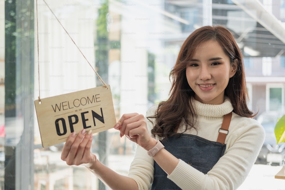 Portrait asian woman smiling and looking to camera in coffee shop. Barista female holding open sign in hand. Working woman small business owner or sme concept.