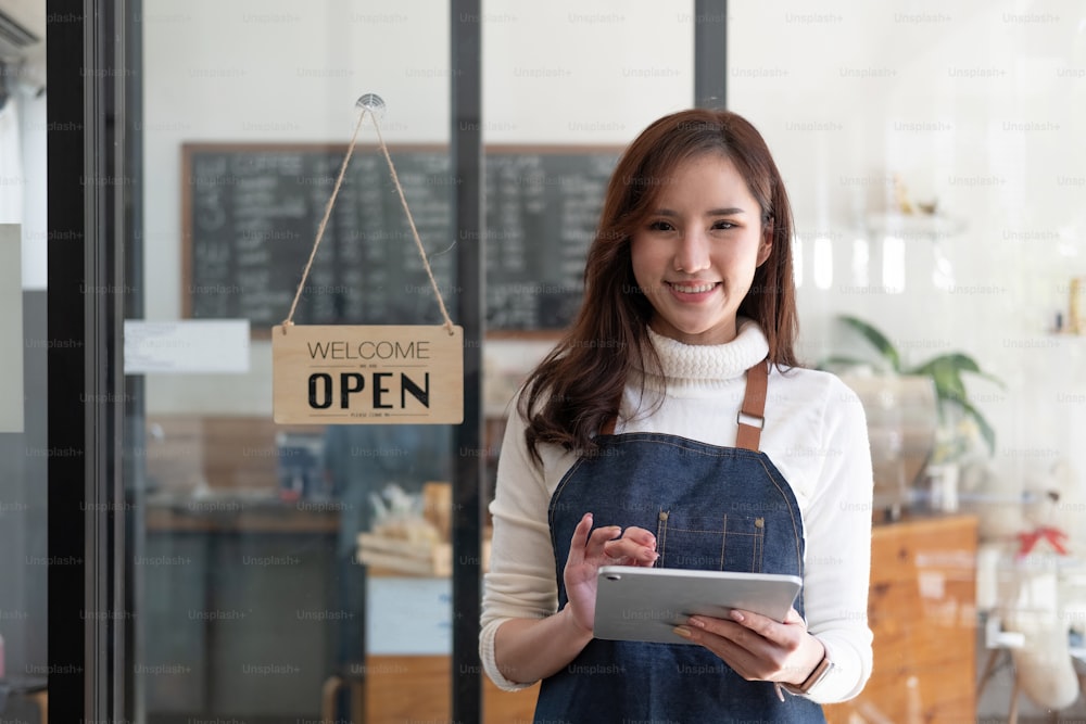 열린 간판이 있는 카페 카운터 뒤에 서 있는 웃는 아시아 기업가의 초상화.