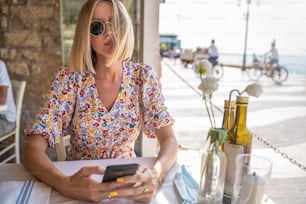 Blonde woman sitting in the restaurant, using mobile phone. Influencer. Summer sunny day. Traveler. Tourist.