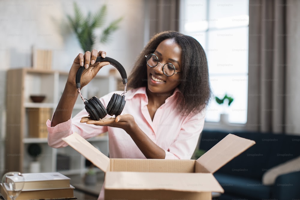 Smiling african woman sitting at table and opening cardboard box with her new wireless headphones. Pretty lady in eyeglasses feeling satisfaction from ordered goods online.