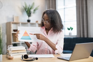 Insegnante femminile intelligente in occhiali che puntano sul diagramma durante la video lezione da casa. Donna afroamericana seduta alla scrivania con laptop moderno e cuffia.