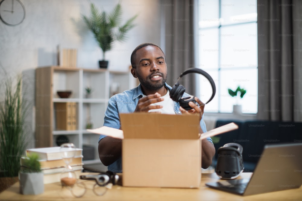 African american man in casual wear taking modern headphones for listening to music from paper box while sitting at the table at home. Concept of technology and blogging.