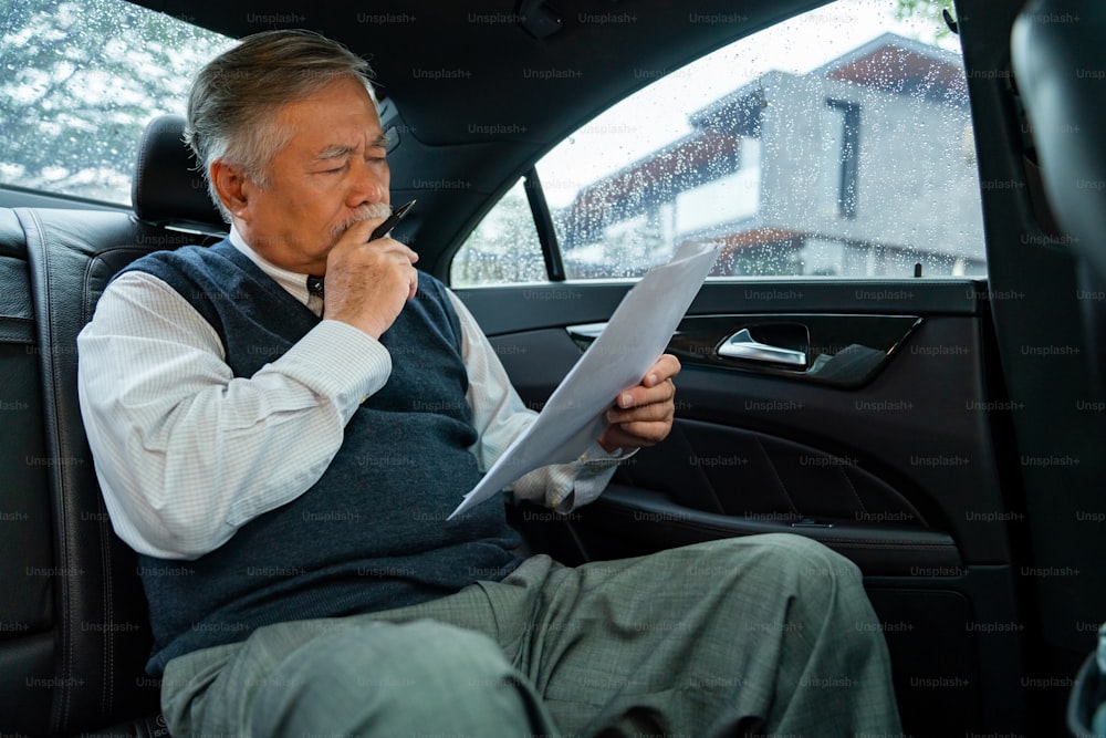 Hombre de negocios asiático de alto nivel con traje sentado en el asiento trasero del automóvil y escribiendo un plan de negocios mientras va a la oficina. Anciano CEO que trabaja en el automóvil y mira el tráfico por la ventana