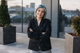 Porträt einer jungen blauhaarigen Geschäftsfrau, die mit gefalteten Händen vor einem Unternehmen steht. Lächelndes Gesicht.
