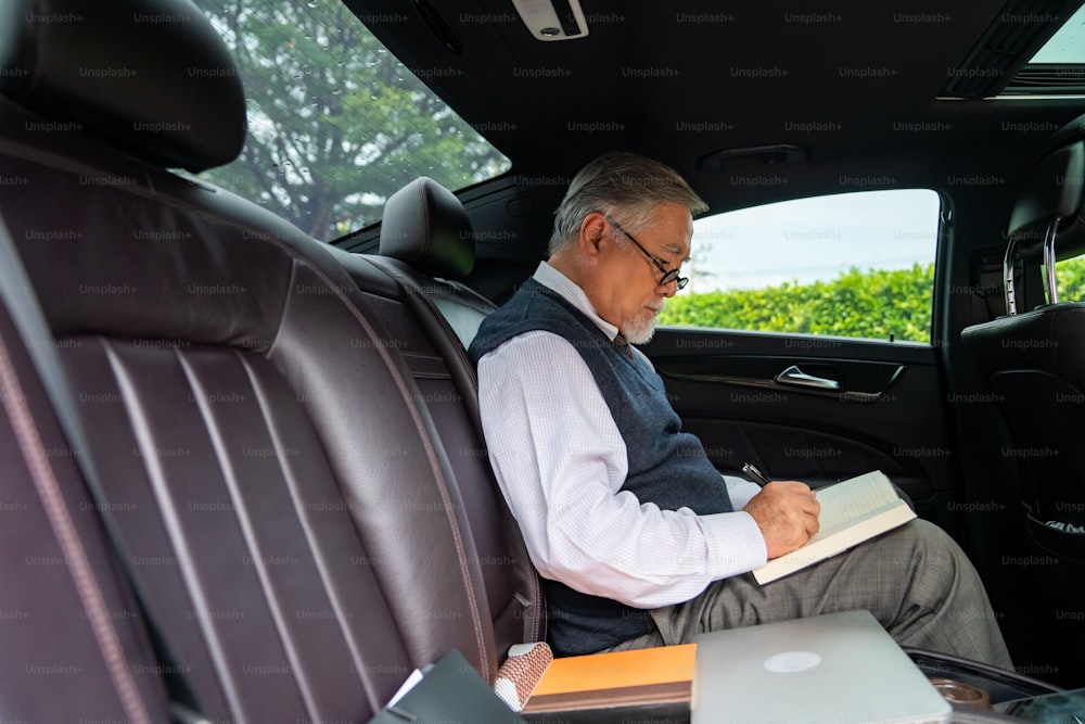 Vertrauen Senior Geschäftsmann CEO im Anzug mit Brille sitzt auf dem Rücksitz des Autos und schreibt Geschäftsplan in das Buch, während er zur Arbeit im Büro geht. Älterer Geschäftsmann und Transportkonzept.