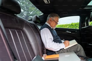 Confiance, homme d’affaires senior, PDG en costume, portant des lunettes, assis sur le siège arrière de la voiture et rédigeant un plan d’affaires dans le livre tout en allant travailler au bureau. Homme d’affaires âgé et concept de transport.