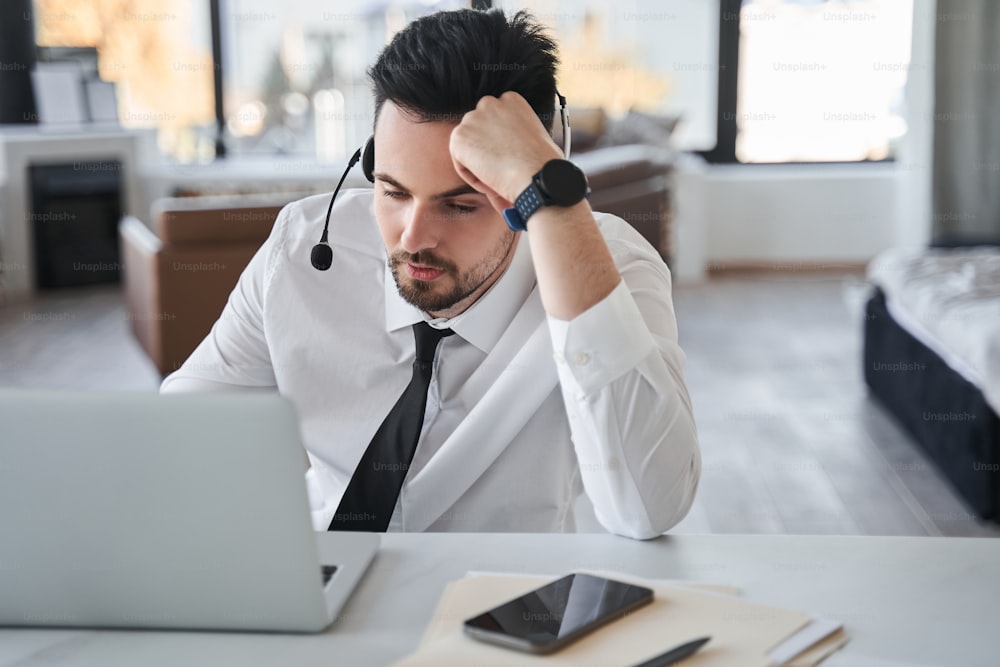 Frustrated operator of the call center wearing special headset looking at the laptop screen and feeling tired while working remotely from home