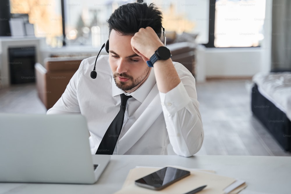 Frustrierter Mitarbeiter des Callcenters, der ein spezielles Headset trägt, auf den Laptop-Bildschirm schaut und sich müde fühlt, wenn er von zu Hause aus arbeitet