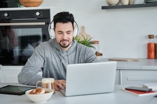 Uomo caucasico che indossa cuffie seduto al tavolo davanti al suo laptop e sorridente mentre effettua una videochiamata con i colleghi