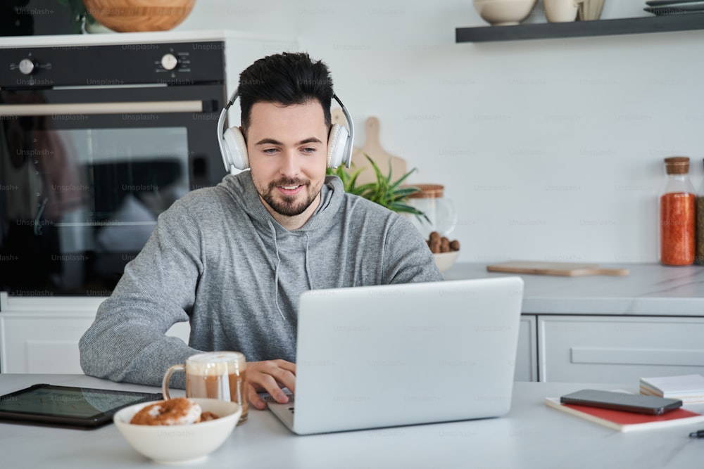 Caucasian man wearing headphones sitting at the table in front of his laptop and smiling while having video call with colleagues