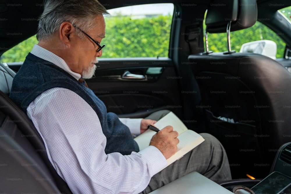 Confidence senior businessman CEO in suit wearing glasses sitting on car back seat and writing business plan in the book while going to work at office. Elderly businessman and transportation concept.