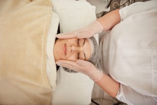 Vue de dessus d’une belle femme allongée sur un lit de repos et souriante tout en recevant un traitement du visage dans une clinique de cosmétologie