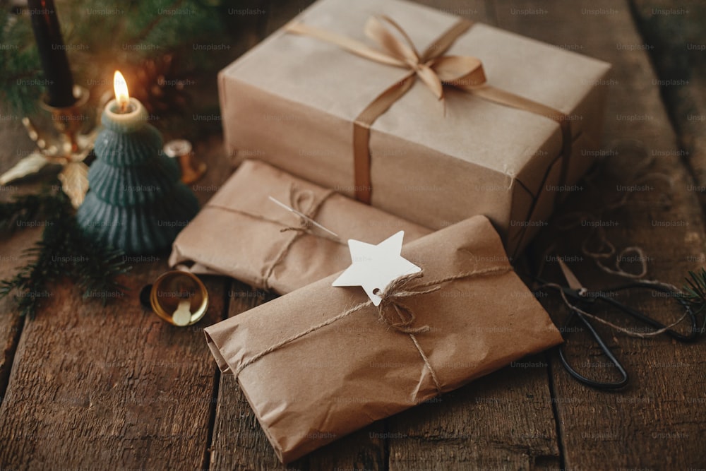 Frohe Weihnachten! Stilvolle Weihnachtsgeschenke eingewickelt in Bastelpapier, Kerze, Schere, Tannenzweige auf rustikalem Holz. Moderne einfache umweltfreundliche Weihnachten präsentiert, atmosphärisch stimmungsvolles Bild.