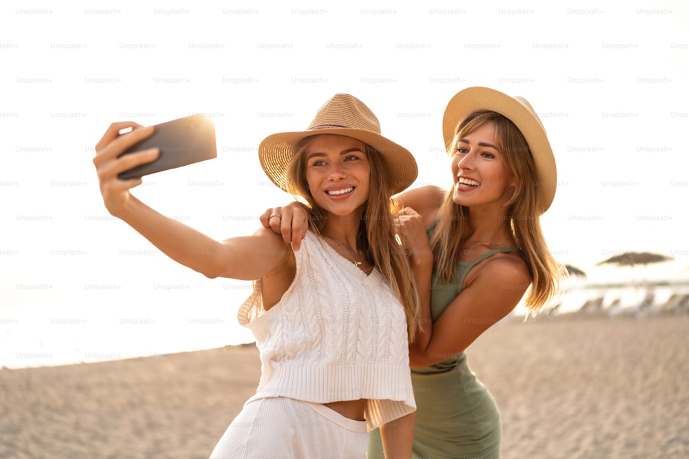 Two happy blonde caucasian sisters having fun on the beach during summer, taking selfie with mobile phone, smiling and looking to the smartphone camera. Beautiful girls wearing hats. Friendship. Real people emotions.