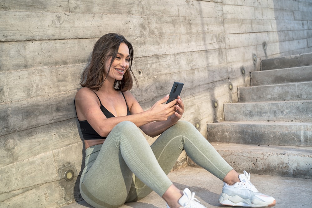 Happy young fit sporty woman in sportswear taking break from morning run, smiling cheerful, talking online by mobile phone or scrolling social media. Healthy lifestyle concept.