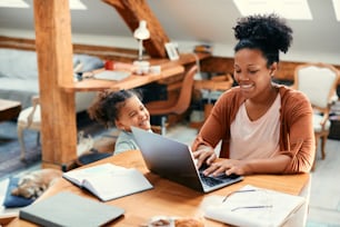 행복한 아프리카계 미국인 딸이 집에서 컴퓨터 작업을 하고 있는 어머니와 이야기하고 있습니다.