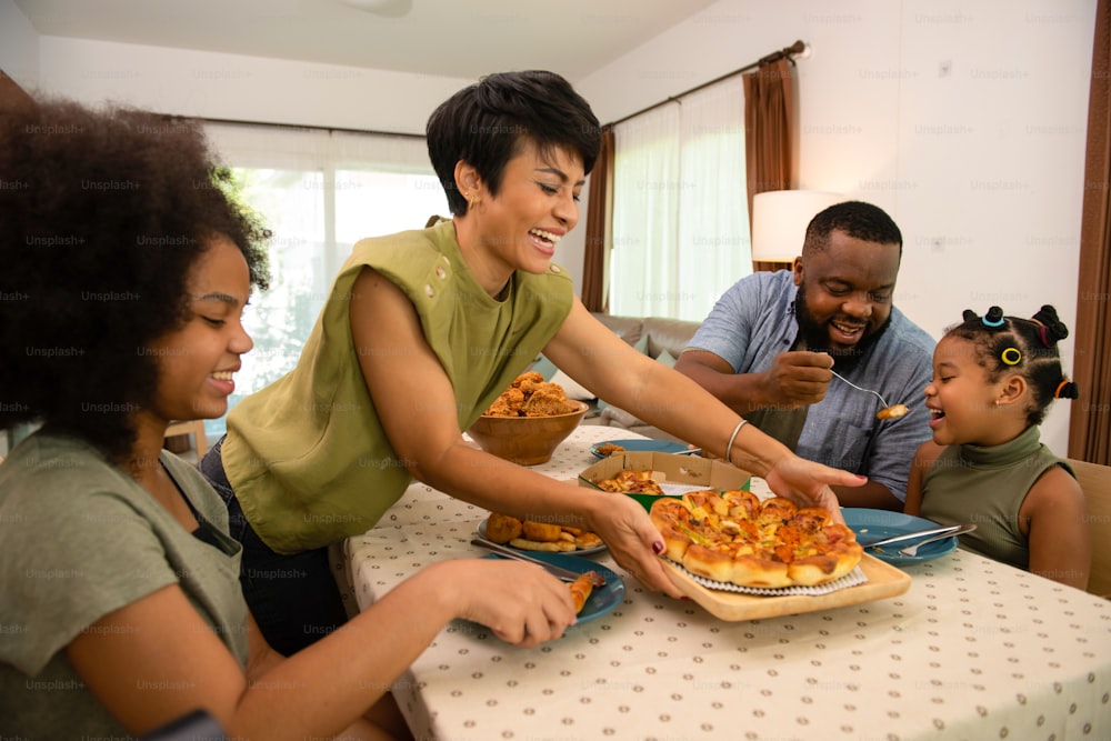 Glückliche afrikanische Familieneltern und zwei kleine Tochter, die zusammen gebratenes Hühnchen und Pizza zum Abendessen essen. Vater und Mutter und süßes Kind Mädchen genießen und Spaß essen und teilen eine Mahlzeit zusammen zu Hause