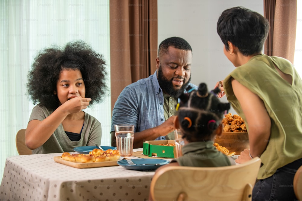 Glückliche afrikanische Familieneltern und zwei kleine Tochter, die zusammen gebratenes Hühnchen und Pizza zum Abendessen essen. Vater und Mutter und süßes Kind Mädchen genießen und Spaß essen und teilen eine Mahlzeit zusammen zu Hause