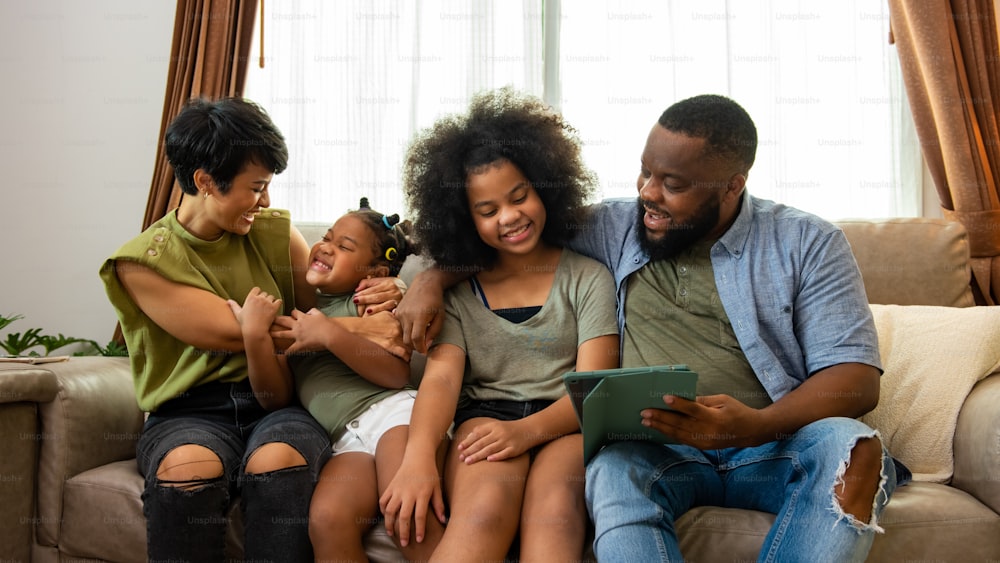 Pais mestiços africanos e duas filhas pequenas sentadas no sofá da sala de estar usando o tablet digital jogam ou assistem a filmes juntos. Família feliz desfruta da atividade de fim de semana com a tecnologia juntos em casa