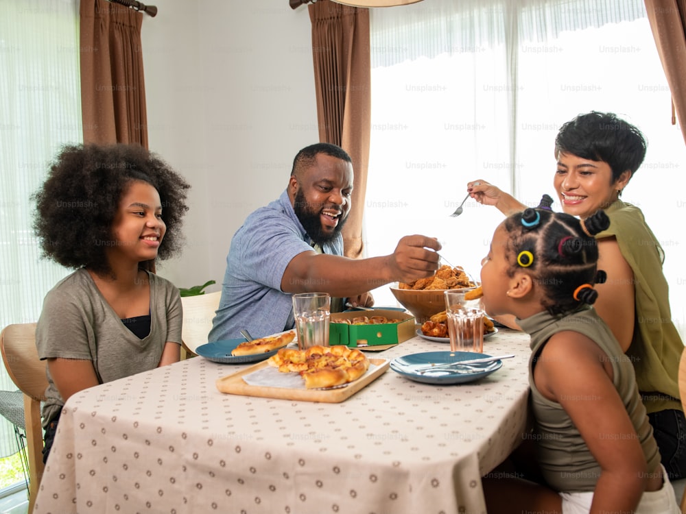 Happy African family parents and two little daughter eating fried chicken and pizza for dinner together. Father and mother and cute child girl kid enjoy and fun eating and sharing a meal together at home