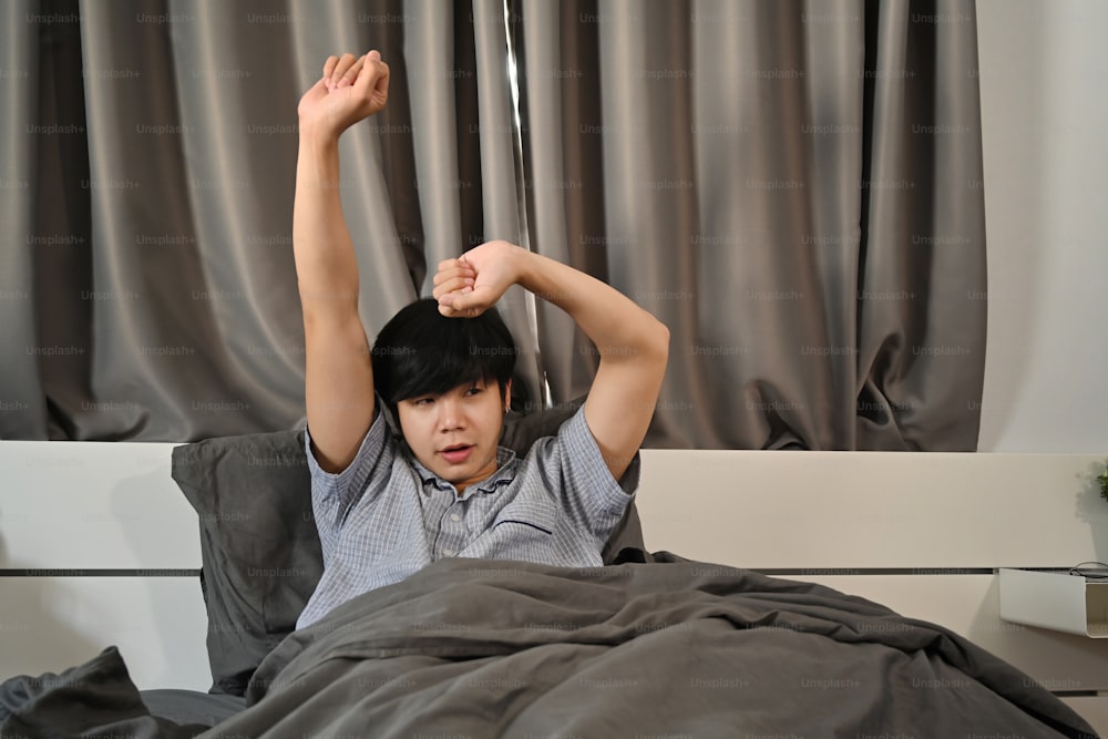 Asian man waking up in bed and stretching his arms.