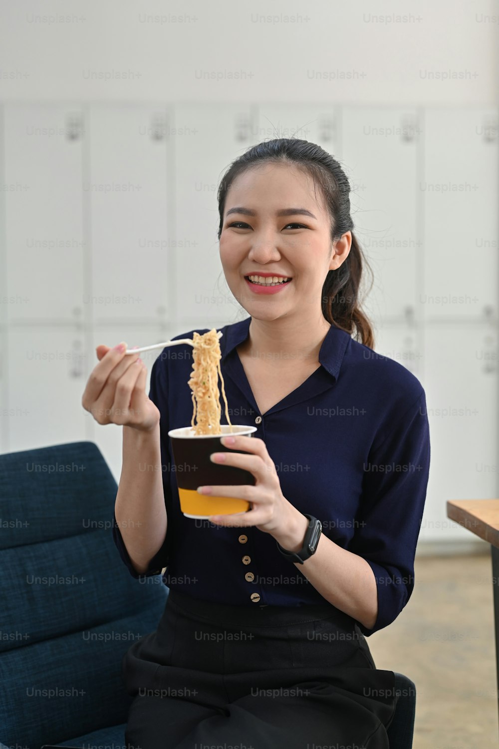 Mulher asiática sorridente segurando garfo de plástico comendo macarrão instantâneo no escritório.